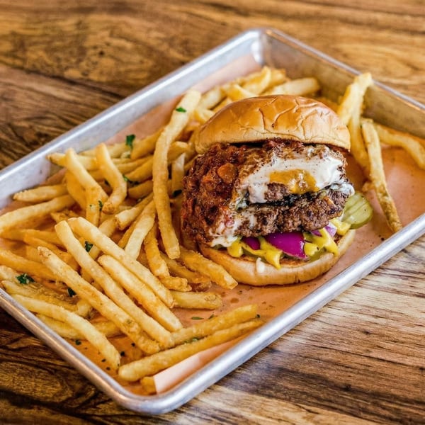 A burger with a fried egg, pickles, and mustard, served with a side of fries on a metal tray.