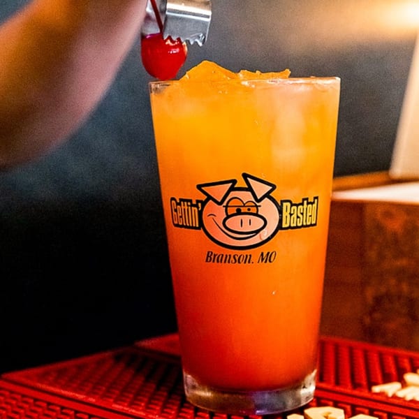 A hand garnishes a tall glass of orange cocktail with a cherry, featuring the Gettin' Basted logo.