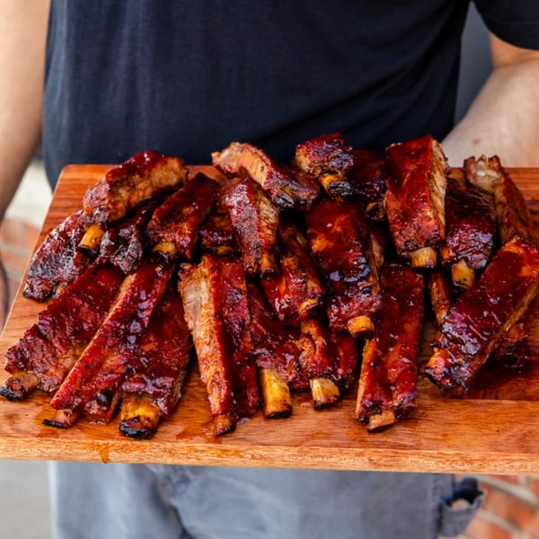 A person holding a wooden board piled high with saucy, glistening Gettin' Basted BBQ ribs.
