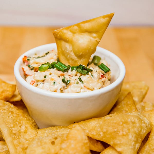 A white bowl of creamy dip with green onions and red peppers, surrounded by crispy wonton chips.