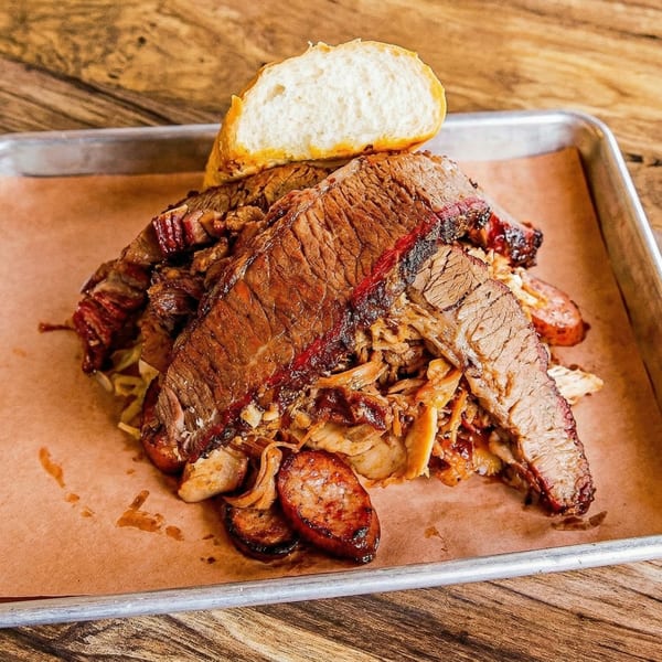 Smoked brisket, pulled pork, and sausage slices with a side of bread on a metal tray.