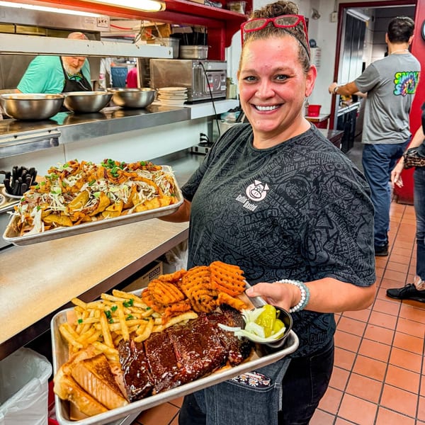 Smiling woman in a restaurant kitchen holding two trays of BBQ ribs, fries, and loaded potato wedges.