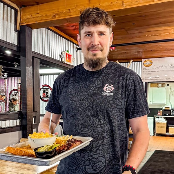 A man in a "Gettin' Basted" t-shirt holds a tray with BBQ, mac and cheese, and fries.