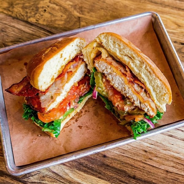 A chicken sandwich, cut in half, with bacon, lettuce, tomato, and pickles on a metal tray.