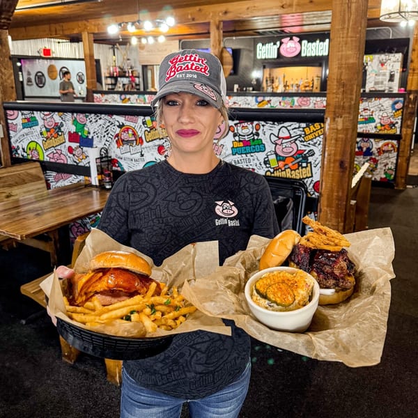A woman in a "Gettin' Basted" hat and shirt holds two baskets of BBQ food.