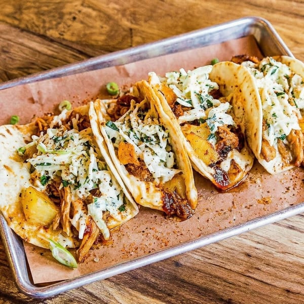 Four loaded tacos with meat, pineapple, and coleslaw on a metal tray with parchment paper.