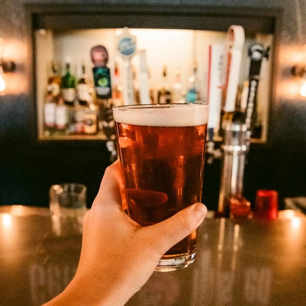 Hand holding a pint of amber beer with a foamy head, in front of a dimly lit bar.