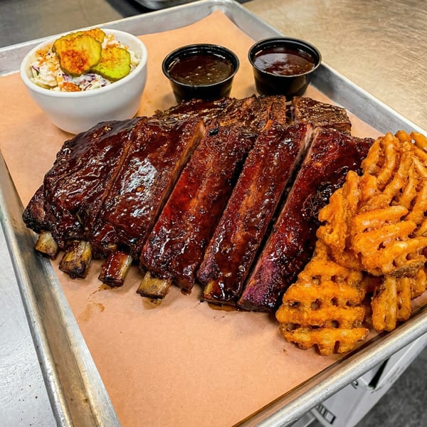 A full rack of BBQ ribs, waffle fries, coleslaw, and two dipping sauces on a metal tray.